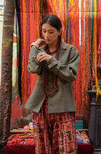 A person wearing a green 'Ananta' jacket with hand embroidery, paired with a patterned outfit, standing in front of a backdrop with colorful fabric strips.