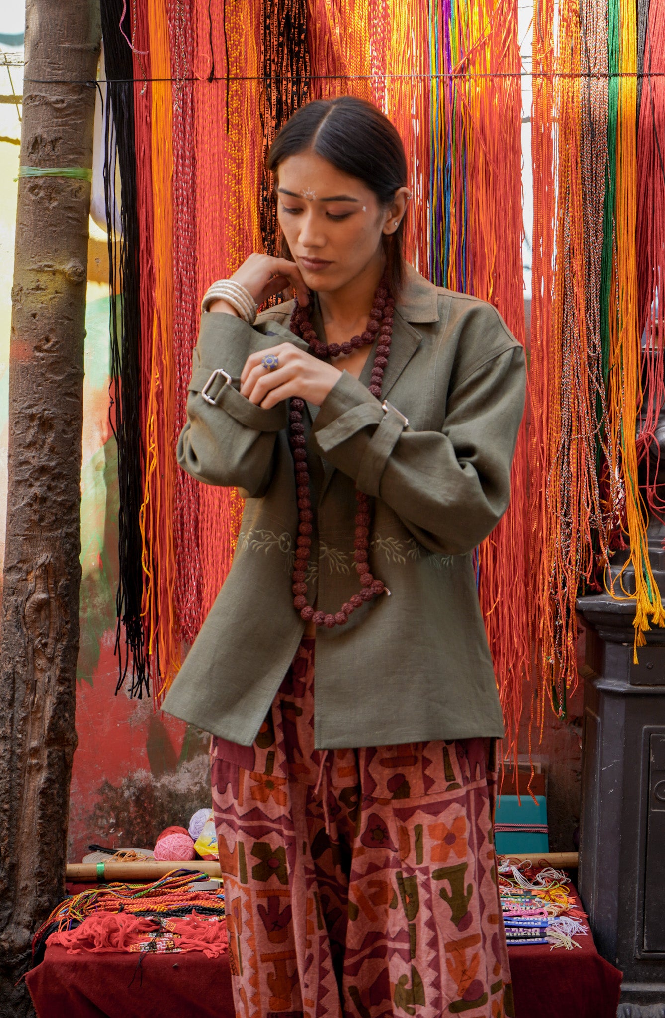 A person wearing a green 'Ananta' jacket with hand embroidery, paired with a patterned outfit, standing in front of a backdrop with colorful fabric strips.